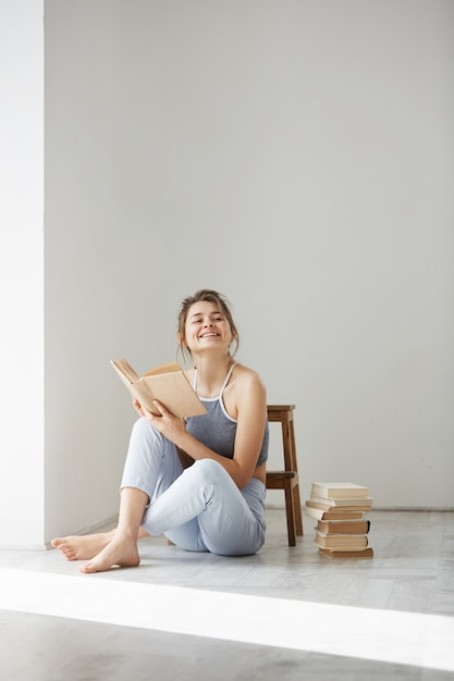 Бесплатное фото Молодая красивая нежная женщина, улыбаясь, держа книгу, сидя на полу на белой стене рано утром.