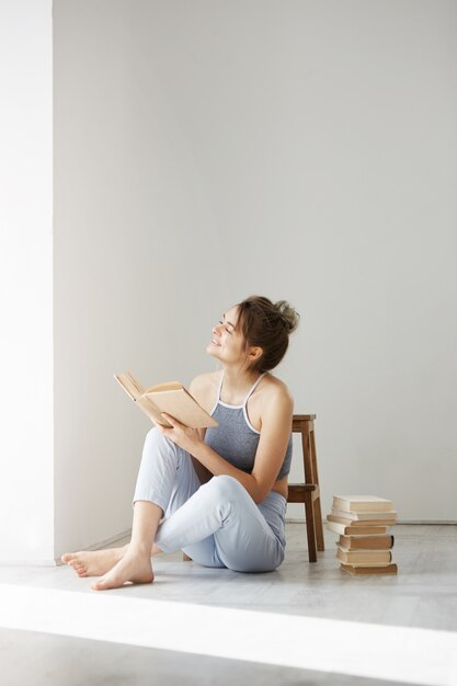 Молодая красивая нежная женщина, улыбаясь, глядя на окно, держа книгу, сидя на полу над белой стеной рано утром.