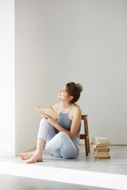 Young beautiful tender woman smiling looking at window holding book sitting on floor over white wall early in morning.