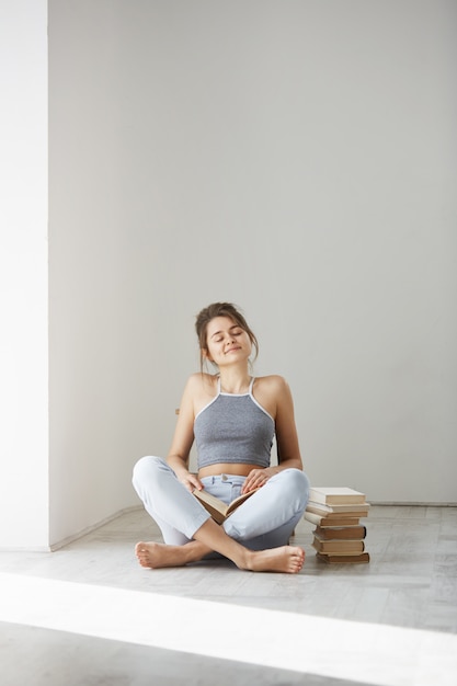 Young beautiful tender woman smiling with closed eyes holding book sitting on floor over white wall early in morning.