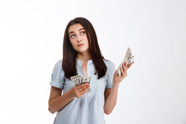 Young beautiful thoughtfully looking up business girl holding money over white wall