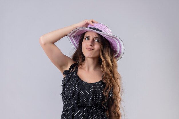 Young beautiful traveler girl in dress in polka dot in summer hat displeased touching her hat nervous and anxious standing over white background