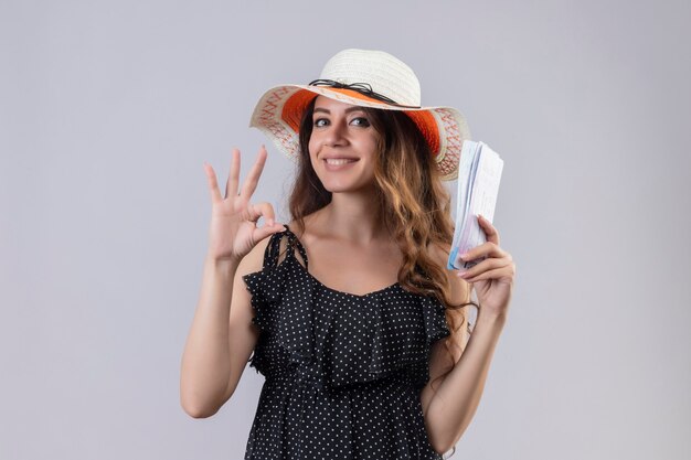 Young beautiful traveler girl in dress in polka dot in summer hat holding air tickets smiling cheerfully doing ok sign standing over white background