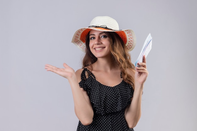 白い背景の上に立っている彼女の手の腕を元気よく提示する笑顔の航空券を保持している夏の帽子の水玉のドレスの若い美しい旅行者の女の子
