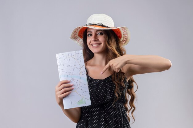 Young beautiful traveler girl in dress in polka dot in summer hat holding map pointing with finger to it smiling cheerfully standing over white background