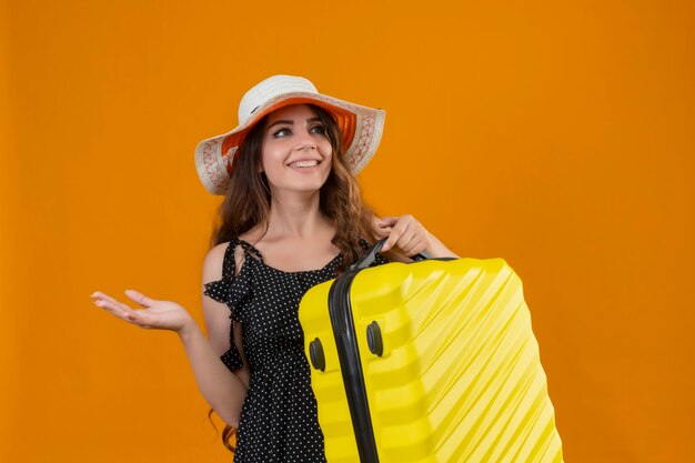 Молодая красивая девушка путешественника в платье в горошек в летней шляпе держит чемодан, глядя в сторону со счастливым лицом, весело улыбаясь, представляя руку, стоящую на желтом фоне