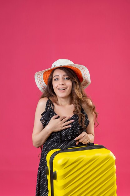 Young beautiful traveler girl in dress in polka dot in summer hat holding suitcase looking at camera smiling cheerfully happy and positive standing over pink background