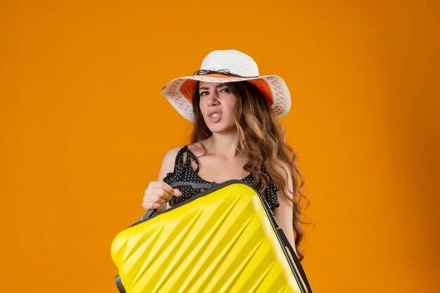 Young beautiful traveler girl in dress in polka dot in summer hat holding suitcase looking at camera with skeptic expression standing over yellow background