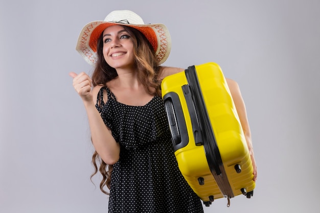 Молодая красивая девушка путешественника в платье в горошек в летней шляпе держит чемодан, выглядящий возбужденным и счастливым, показывает палец вверх, радуясь ее успеху и победе, стоя на белом фоне
