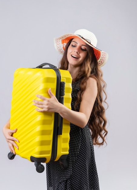 Young beautiful traveler girl in dress in polka dot in summer hat holding suitcase looking at it with happy smile on face standing over white background