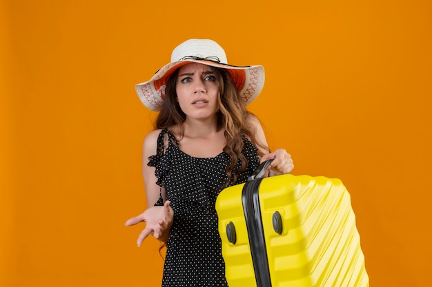 Молодая красивая девушка путешественника в платье в горошек в летней шляпе держит чемодан, нервно и тревожно глядя на желтом фоне