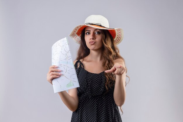 흰색 배경 위에 서있는 얼굴에 회의적인 표정으로 카메라를 손가락으로 가리키는 여름 모자에 폴카 도트 드레스에 젊은 아름 다운 여행자 소녀