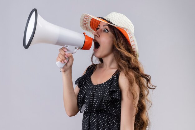 Young beautiful traveler girl in dress in polka dot in summer hat shouting to megaphone surprised and shocked standing over white background