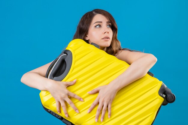 Young beautiful traveler girl holding suitcase nervous and very anxious looking away standing over blue background