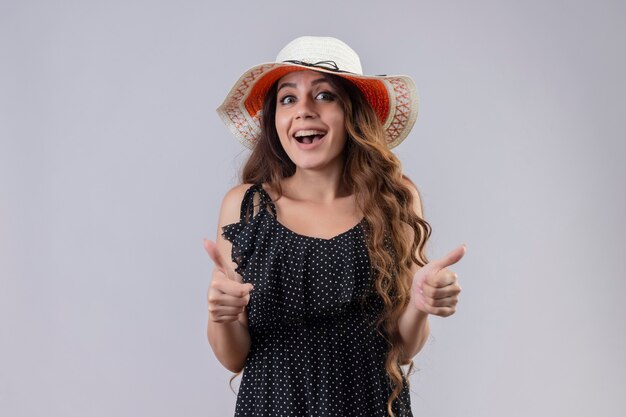 Young beautiful traveler girl in summer hat looking at camera happy and positive smiling cheerfully showing thumbs up standing over white background