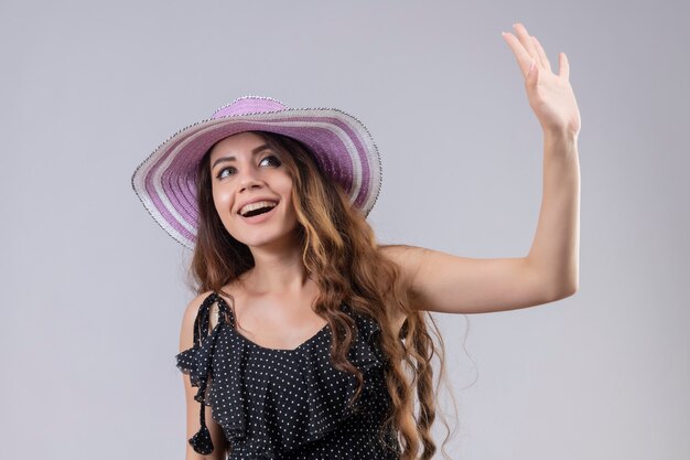 Молодая красивая девушка путешественника в летней шляпе, весело улыбаясь, со счастливым лицом, машет рукой, стоя на белом фоне