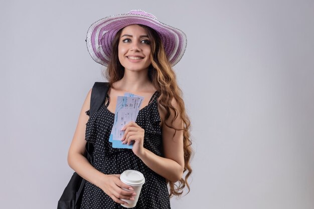 Young beautiful traveler girl in summer hat with backpack holding air tickets smiling cheerfully happy and positive standing over white background