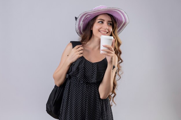 커피 컵을 들고 배낭 여름 모자에 젊은 아름 다운 여행자 소녀 유쾌 하 게 행복 하 고 긍정적 인 흰색 배경 위에 서 멀리 서보고 웃 고