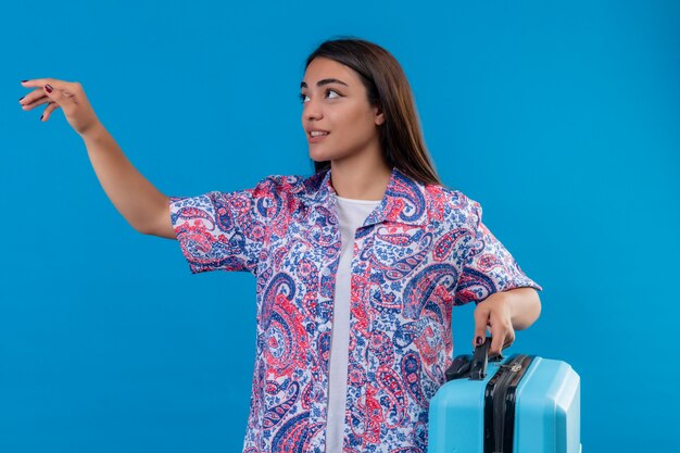 Молодая красивая женщина путешественника держа голубой чемодан смотря в сторону показывать с рукой прося прийти над голубой стеной