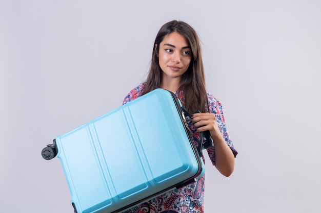 Free photo young beautiful traveler woman holding blue suitcase looking aside with confident smile standing over white background