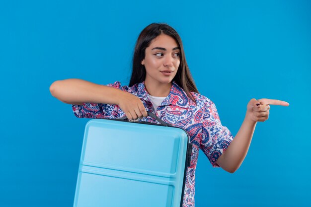 Молодая красивая женщина путешественник держит синий чемодан, указывая пальцем в сторону, уверенно глядя на синюю стену