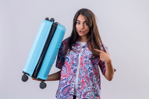 Young beautiful traveler woman holding blue suitcase pointing with index finger to it smiling confident standing over white background