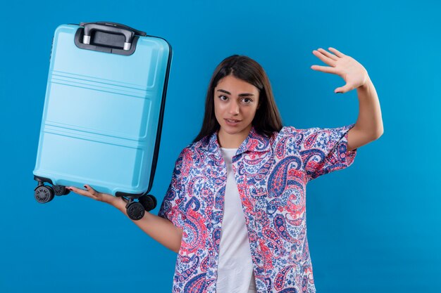 Young beautiful traveler woman holding blue suitcase raising hand in surrender with fear expression over blue wall