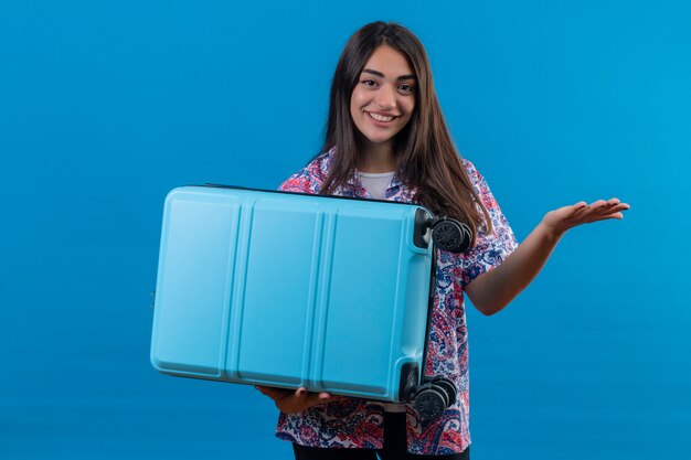 Young beautiful traveler woman holding blue suitcase with happy smile raising hand over blue wall