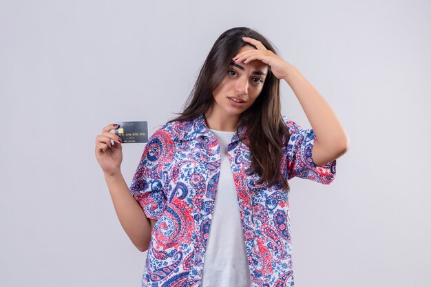 Young beautiful traveler woman holding credit card looking confused touching head , doubtful expression over white wall