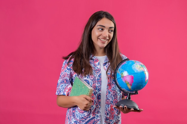 Young beautiful traveler woman holding notebook and globe looking aside with happy face smiling over isolated pink wall