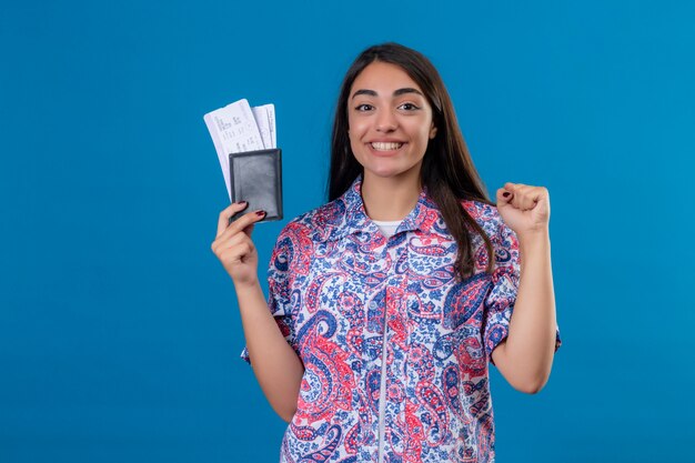 Молодой красивый путешественник женщина держит паспорт с билетами, весело улыбаясь, поднимая кулак после победы, готов к празднику над изолированной синей стеной