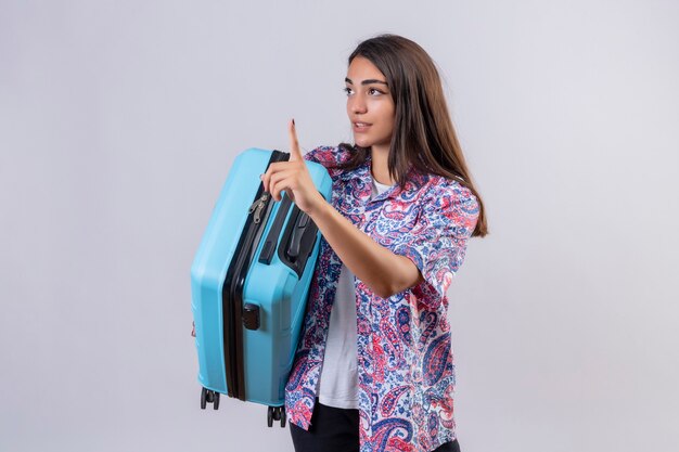 Young beautiful traveler woman holding suitcase gesturing wait a minute with serious confident expression on face standing over white background