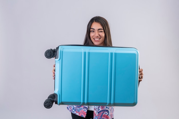 Young beautiful traveler woman holding suitcase looking at camera winking smiling cheerfully standing over isolated white background