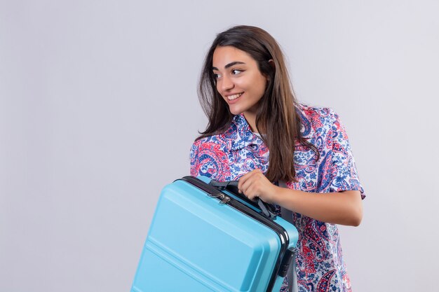 Молодой красивый путешественник женщина держит чемодан, глядя уверенно, позитивно и счастливо, весело улыбаясь, готов путешествовать по белой стене