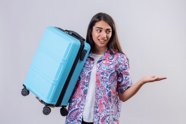 Young beautiful traveler woman holding suitcase looking confused gesturing with hand and expression as asking a question over white wall