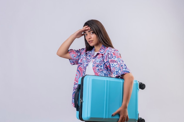 Молодая красивая путешественница женщина, держащая чемодан, выглядит усталой трогательной головой, стоящей на изолированном белом фоне