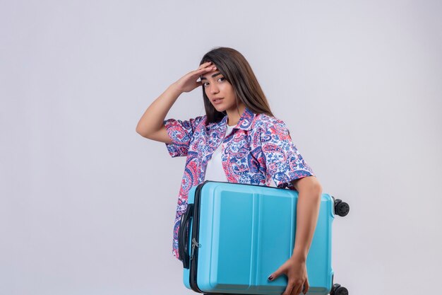 Young beautiful traveler woman holding suitcase looking tired touching head standing over isolated white background