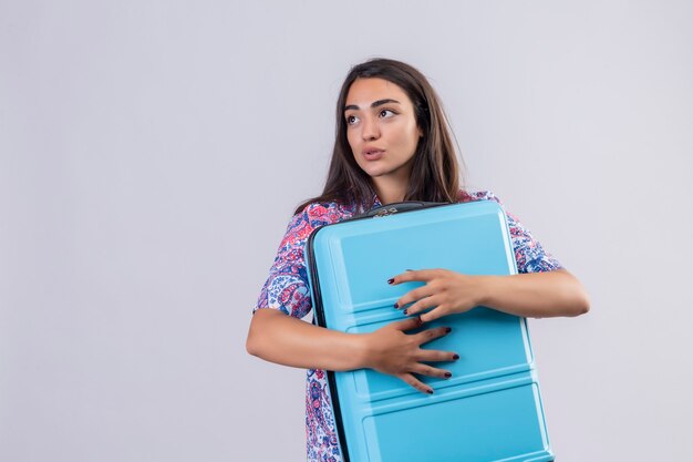 Young beautiful traveler woman hugging blue suitcase looking aside with positive expression on face standing over white background