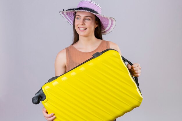 Young beautiful traveler woman in summer hat holding yellow suitcase smiling with happy face standing over white background