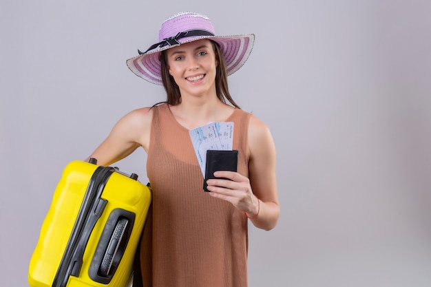 Young beautiful traveler woman in summer hat holding yellow suitcase and tickets smiling with happy face over white wall