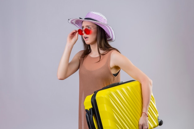 노란색 가방을 들고 빨간 선글라스를 쓰고 여름 모자에 젊은 아름 다운 여행자 여자는 흰색 벽을보고 놀라게 실망