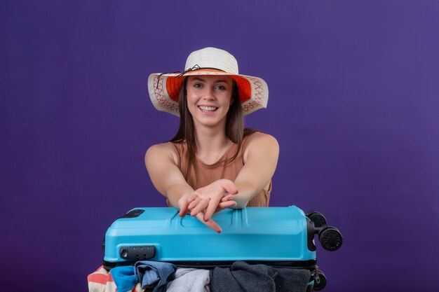 Молодая красивая женщина-путешественница в летней шляпе с чемоданом, полным одежды, оптимистично и счастливо смотрит в камеру с улыбкой на лице, стоя на фиолетовом фоне