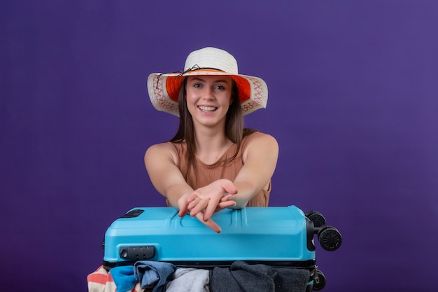 Donna giovane bella viaggiatore in cappello estivo con la valigia piena di vestiti ottimista e felice con il sorriso sul viso sul muro viola