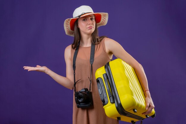 노란색 가방과 카메라가 보라색 배경 위에 서있는 대답이없는 혼란 찾고 여름 모자에 젊은 아름 다운 여행자 여자