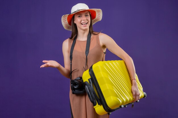 La giovane bella donna del viaggiatore in cappello dell'estate con la valigia gialla e la macchina fotografica sorridenti e felici sorridono allegramente con il braccio si sono alzate sopra la parete porpora