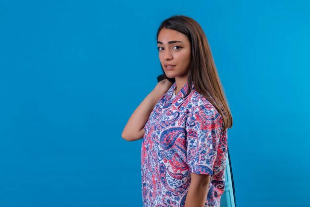 Young beautiful traveler woman with blue suitcase looking uncertain and confused over isolated blue wall