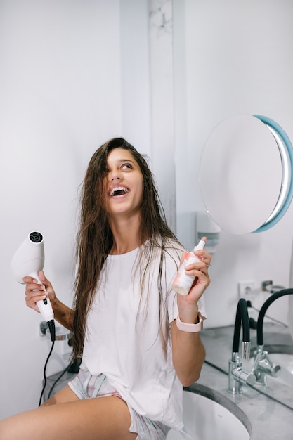 Young beautiful woman in the bathroom holding a hairdryer and a small bottle
