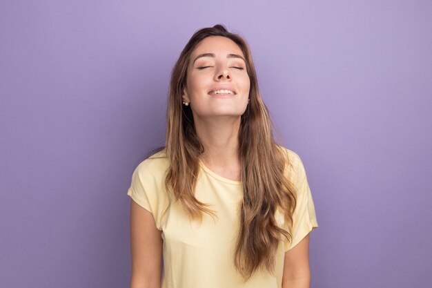 Молодая красивая женщина в бежевой футболке счастлива и позитивно улыбается с закрытыми глазами