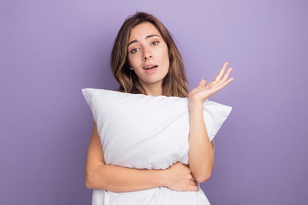 Young beautiful woman in beige t-shirt holding white pillow looking at camera with confident expression with arm raised standing over purple background