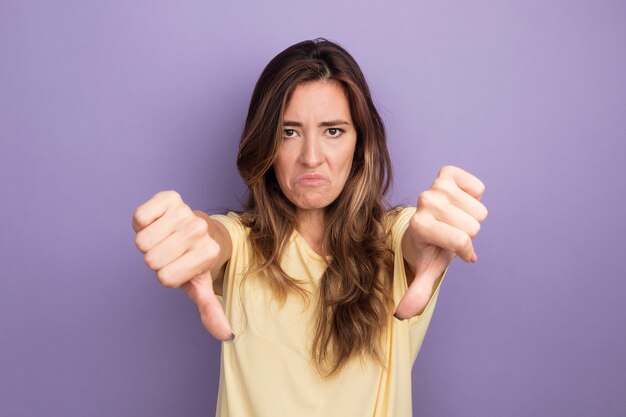 Young beautiful woman in beige t-shirt looking at camera displeased showing thumbs down standing over purple